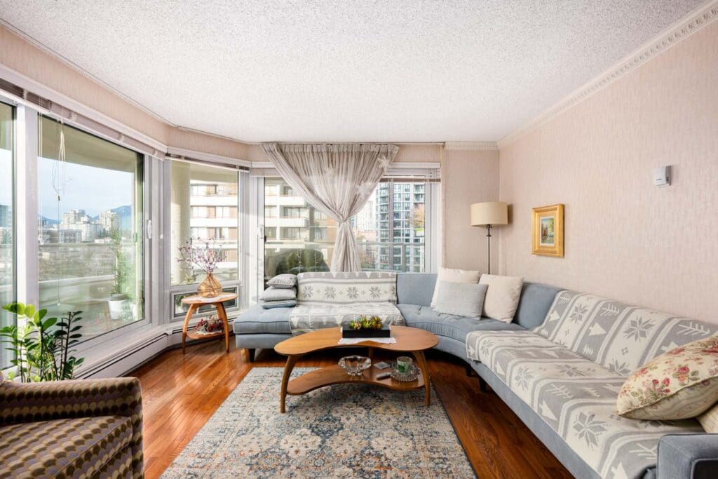 Living room with a large sectional sofa, wooden coffee table, and a patterned rug. Large windows offer a city view. Plants and framed artwork complete the decor.