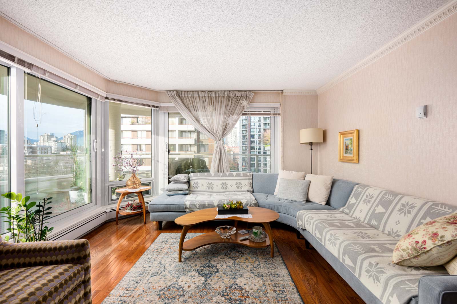 Living room with a large sectional sofa, wooden coffee table, and a patterned rug. Large windows offer a city view. Plants and framed artwork complete the decor.