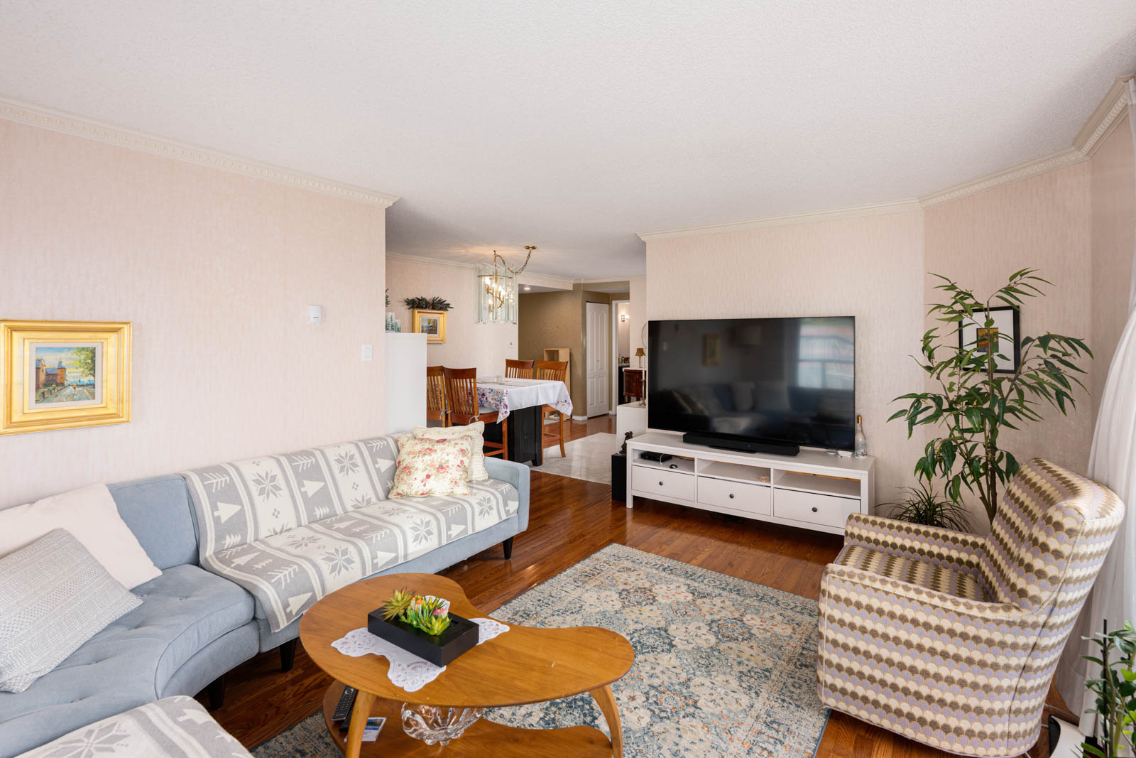 Living room with a patterned couch and armchair, large flat-screen TV on a white stand, rug, wooden coffee table with plant, and dining area in the background.