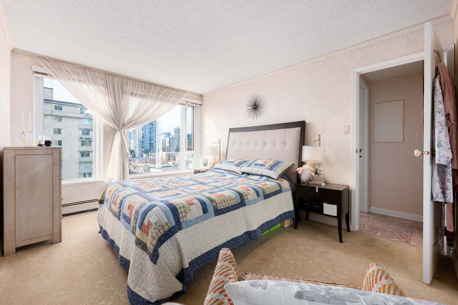 A bedroom with a large bed covered in a patchwork quilt, bedside tables with lamps, a dresser, and two large windows showing a cityscape view.