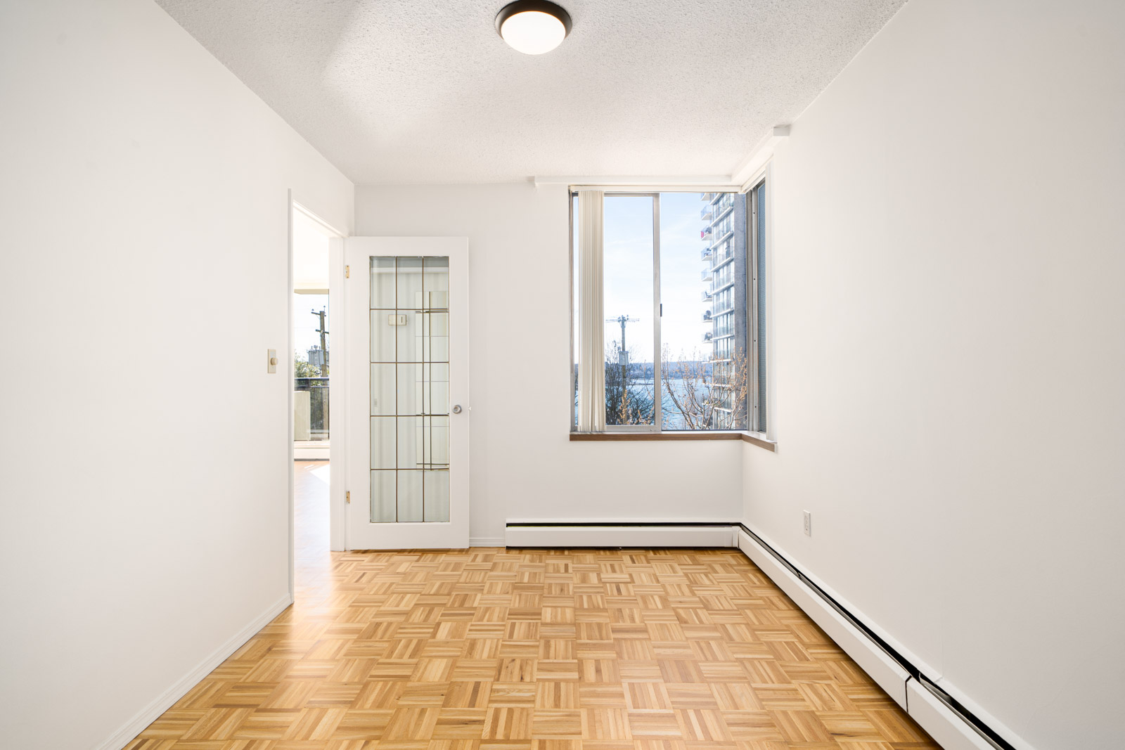 Empty room with parquet floor, a window with a view of a building and water, a glass panel door leading to another room, and a ceiling light fixture.