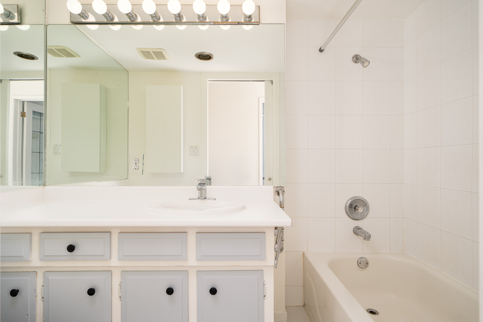 A bathroom with a white sink on a counter, a large mirror with light bulbs above, and a tiled bathtub with a showerhead.