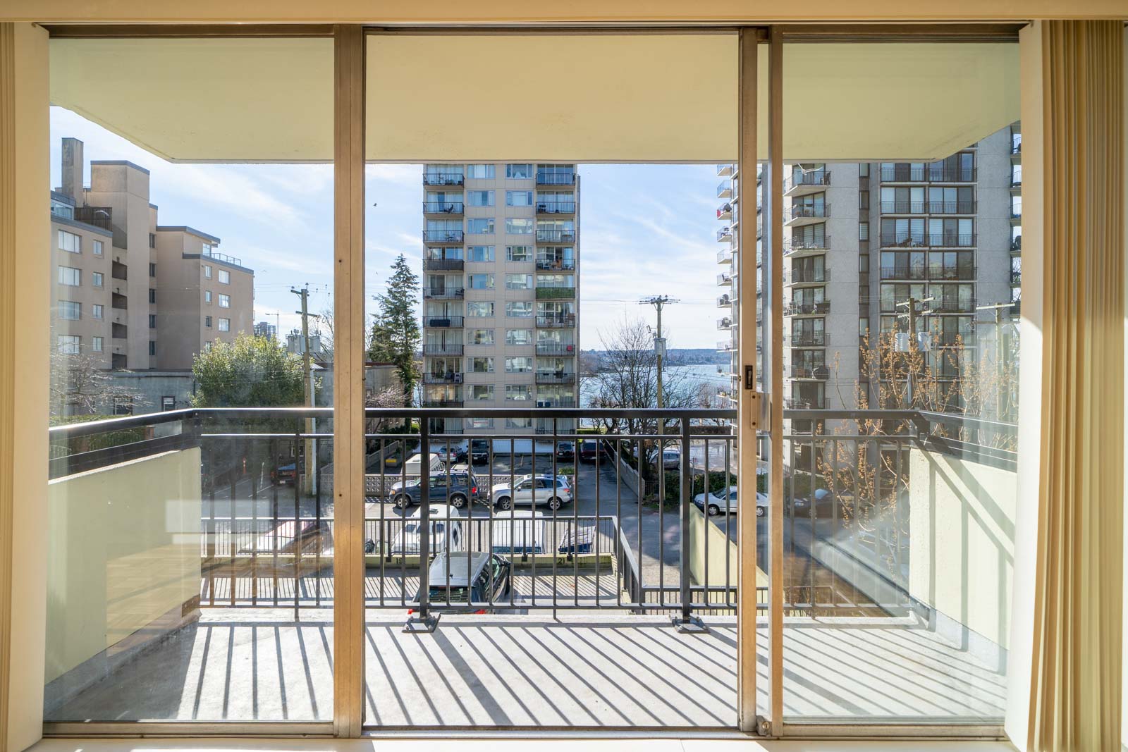 A view through sliding glass doors onto a balcony overlooking a street with several tall buildings and a glimpse of water in the distance.