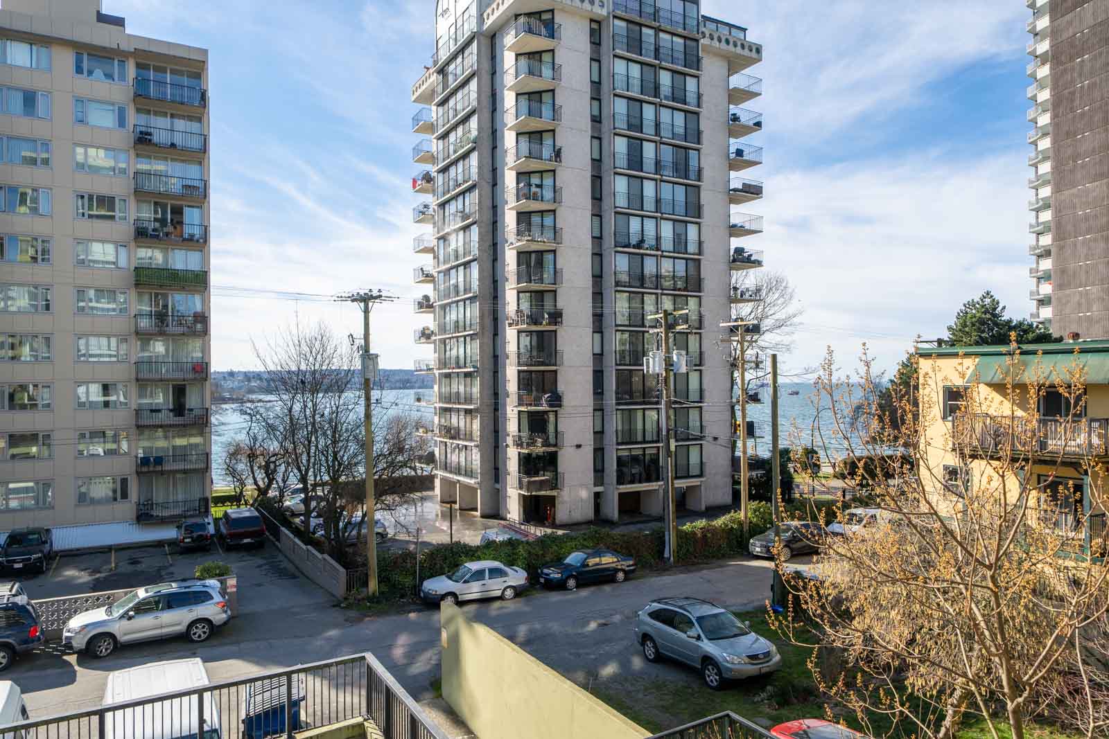 View of urban residential area with several multi-story buildings, parked cars, and a glimpse of the ocean in the background on a clear day.
