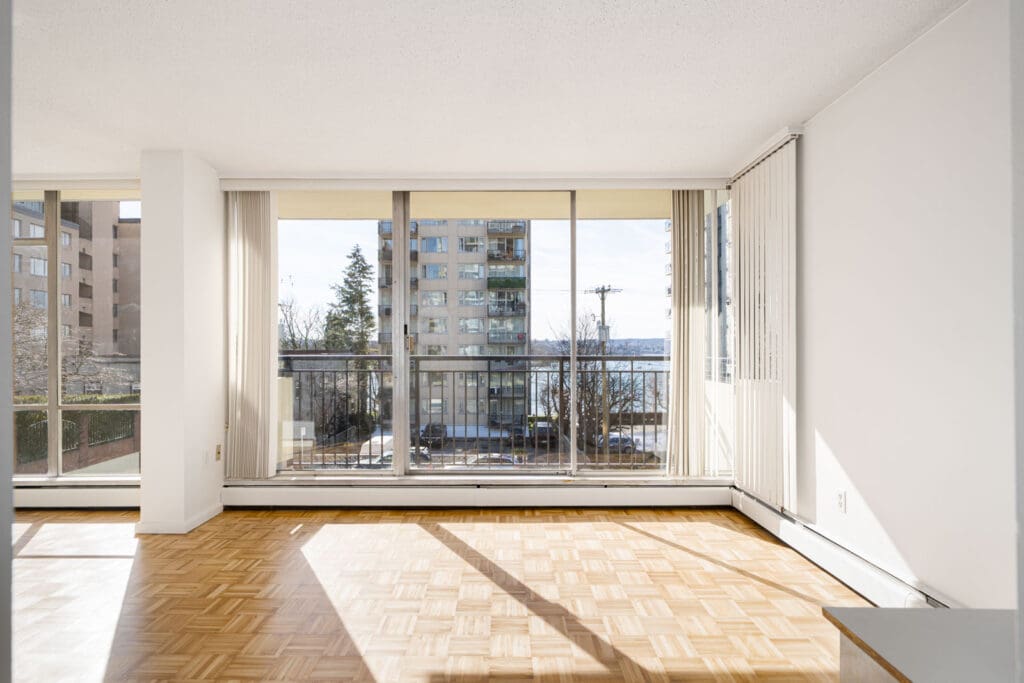 Empty room with large windows, parquet flooring, and a view of urban buildings and trees outside.
