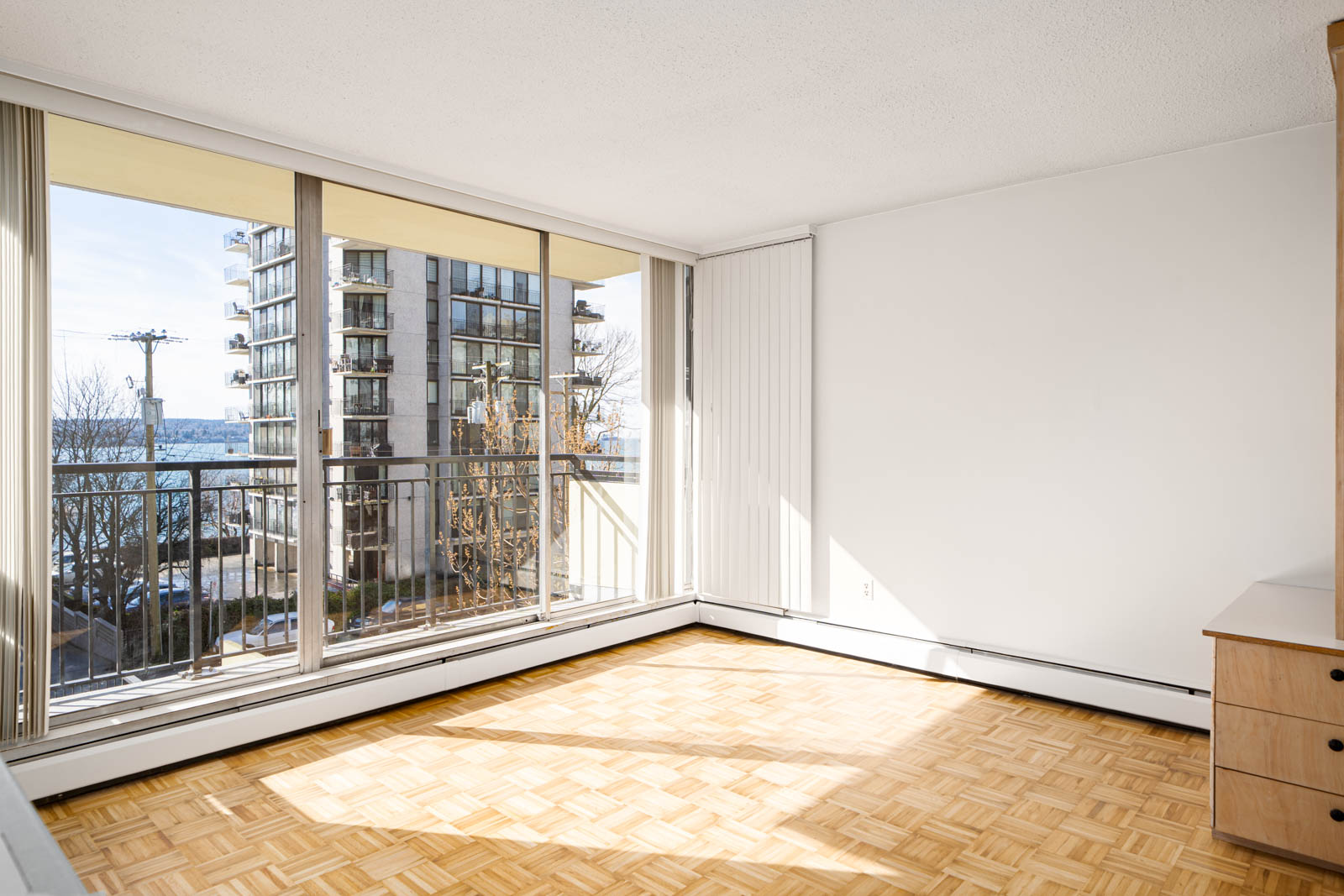Bright room with parquet flooring, large sliding glass doors, and a balcony. Visible buildings and partial ocean view outside.