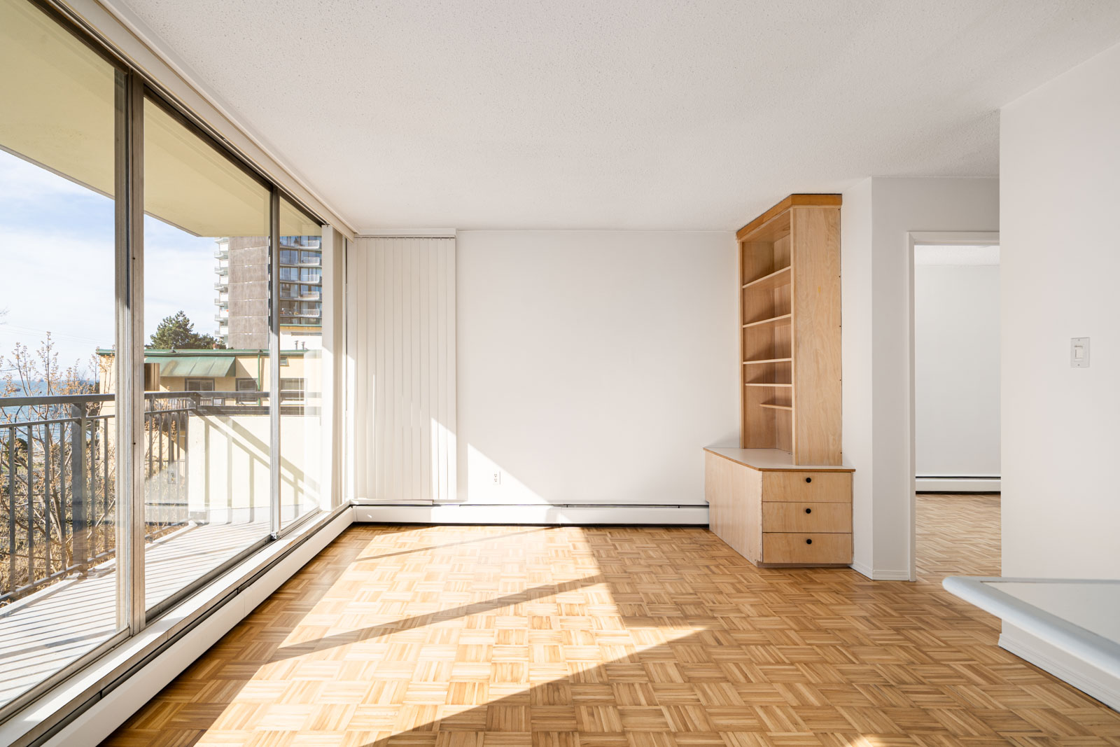 Bright, empty living room with parquet flooring, large sliding glass doors leading to a balcony, and built-in wooden shelving on the right.