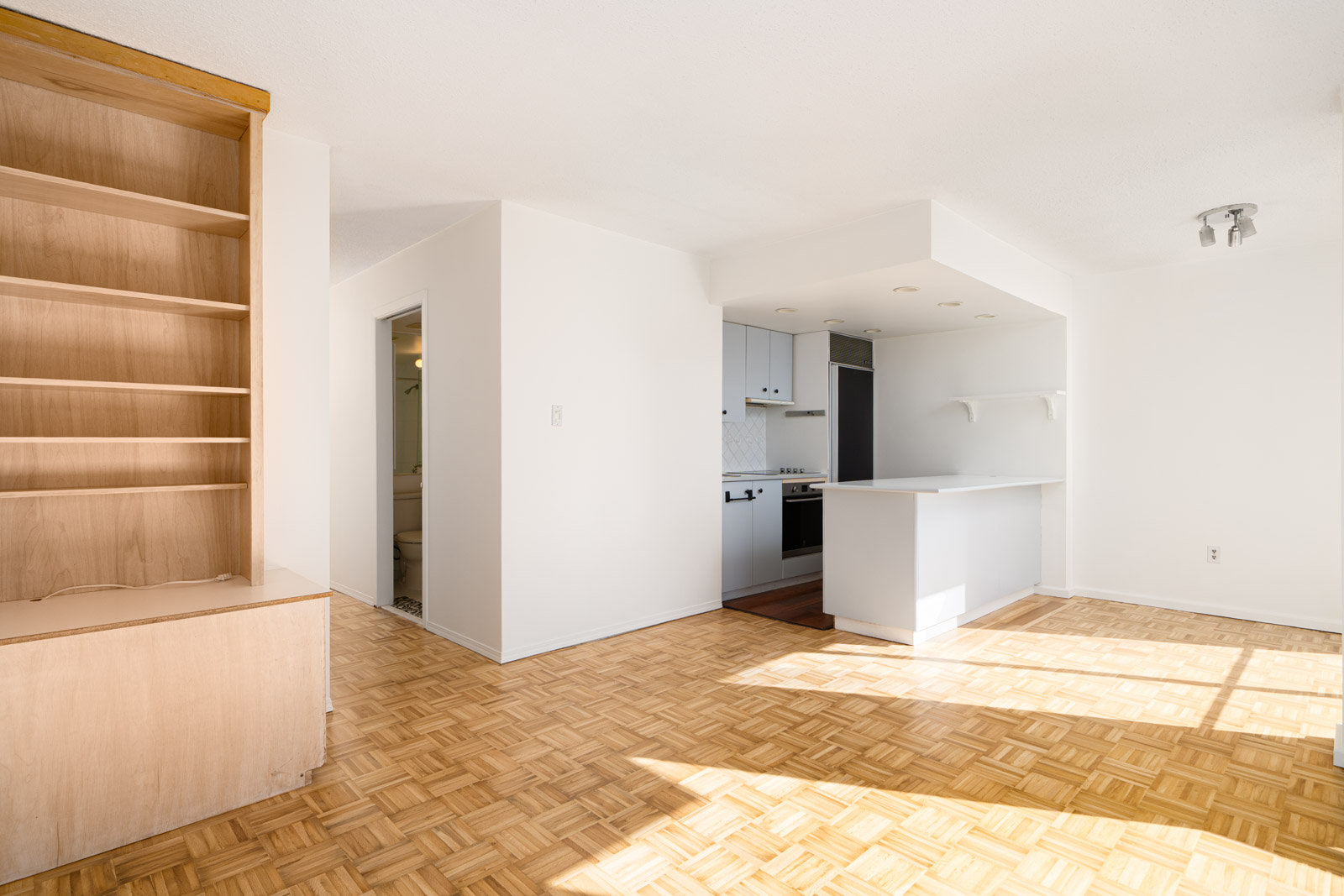 Bright, empty apartment with light wood parquet flooring, built-in shelving, and a small kitchen featuring white cabinets and a counter.