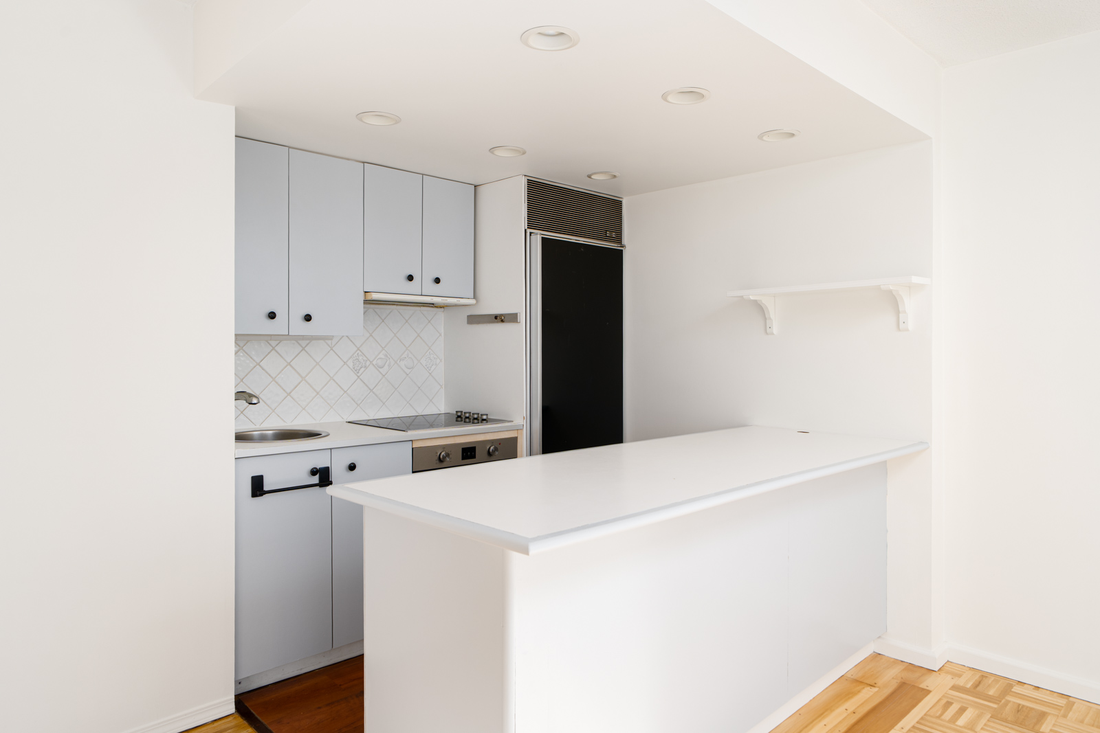 Minimalist white kitchen with a small counter, white cabinets, black refrigerator, and parquet flooring.