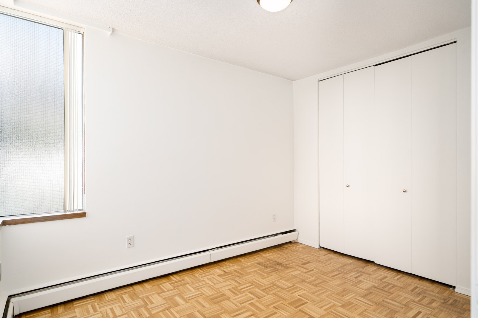 Empty room with white walls, a small window on the left, closed white closet doors on the right, and a light wooden parquet floor.