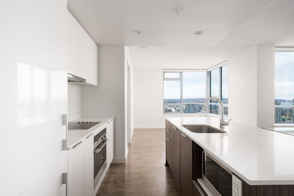 Modern kitchen with sleek white cabinets, built-in oven, and an island with a sink. Large windows provide ample natural light and city views. Brown wooden flooring contrasts the white decor.