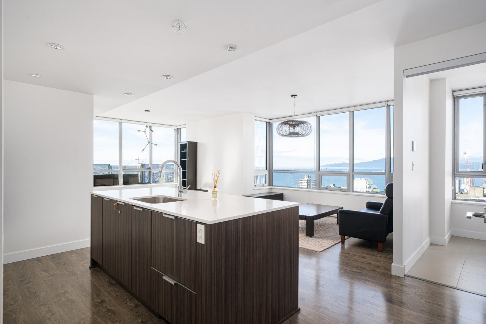 Modern apartment interior featuring a kitchen island, wooden flooring, and large windows with city and ocean views. A seating area includes a sofa and armchair.