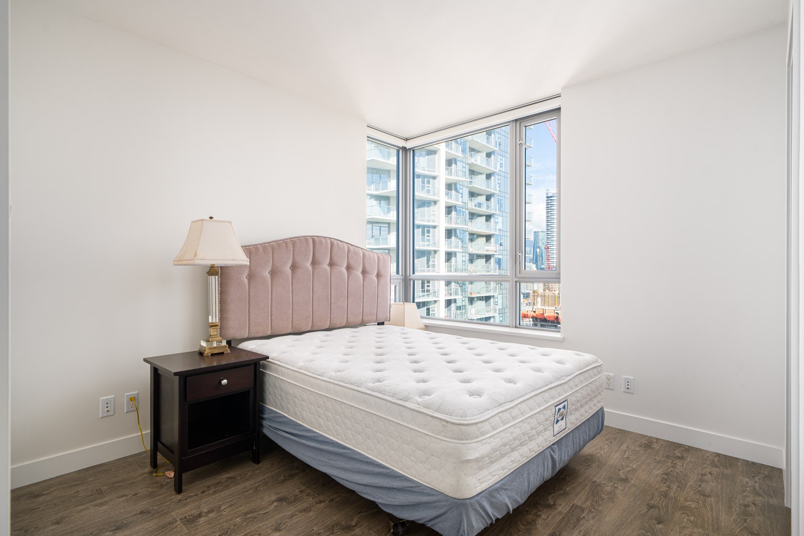 Minimalist bedroom with a bare mattress on a bed frame, a bedside table with a lamp, and large windows displaying a cityscape view.