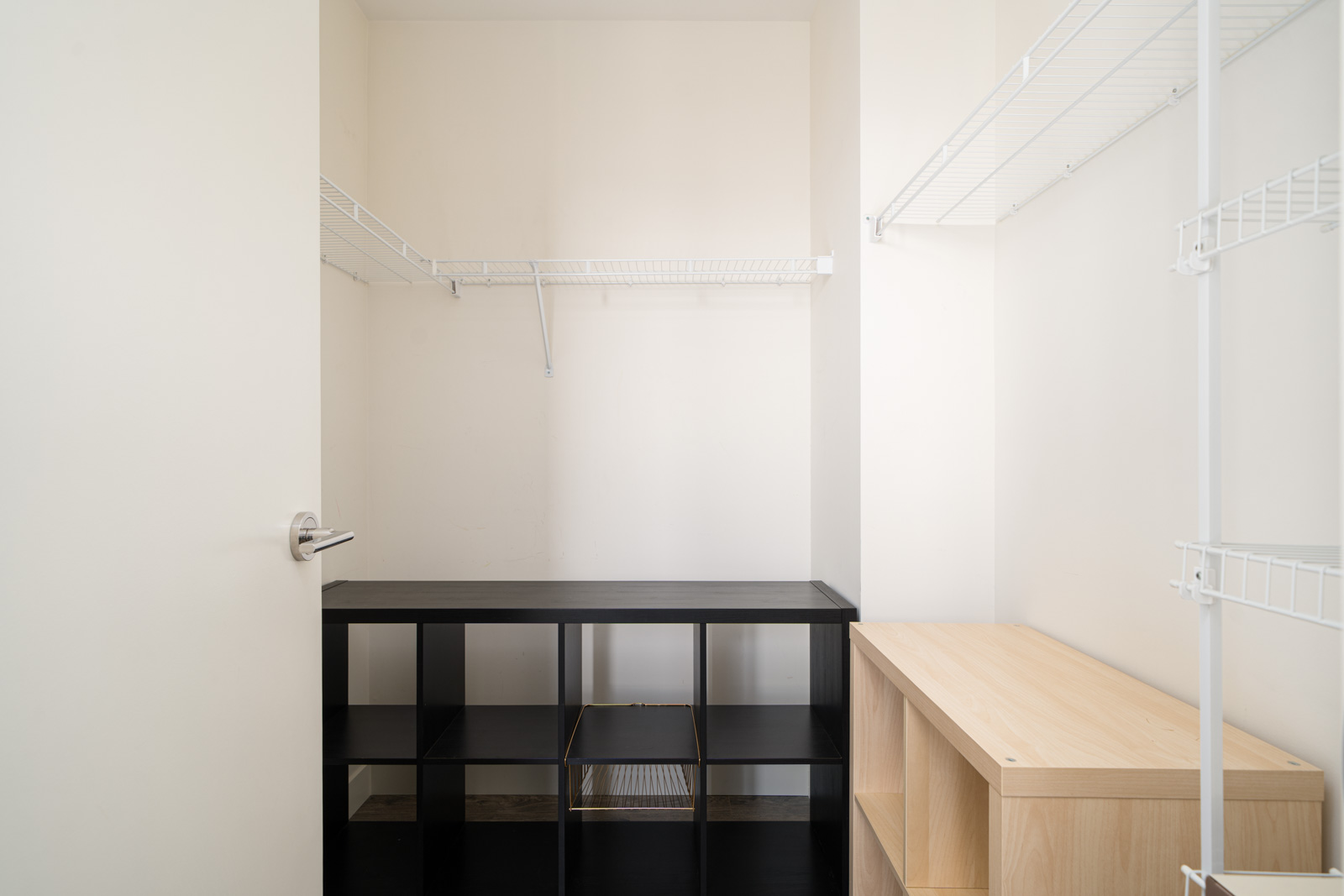 Empty walk-in closet with white walls, featuring black and wood shelves and white wire shelving units on the walls.