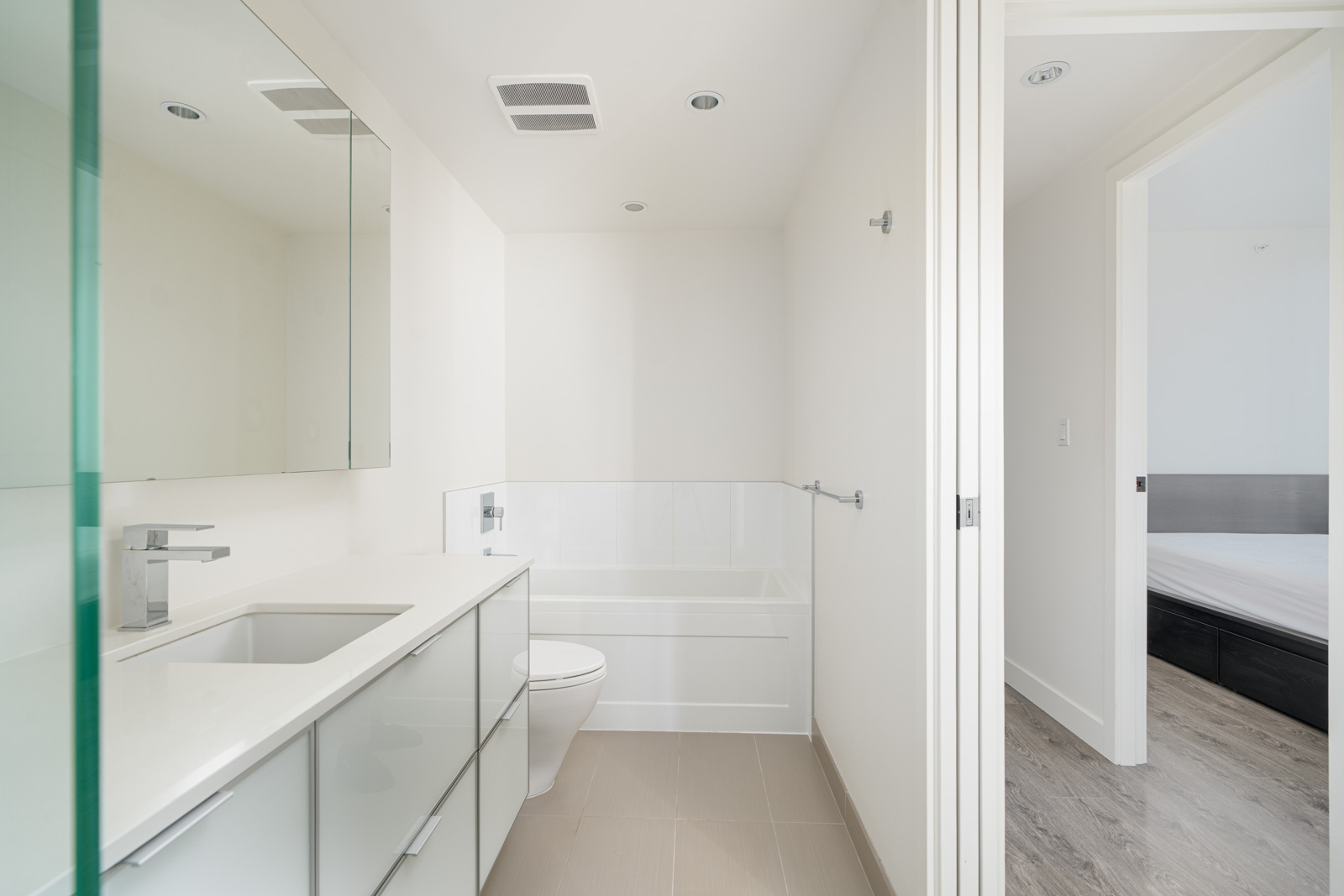 Modern bathroom with a large mirror, white countertop, sink, and bathtub. There's a wall-mounted toilet and open door leading to a bedroom with a wooden floor.