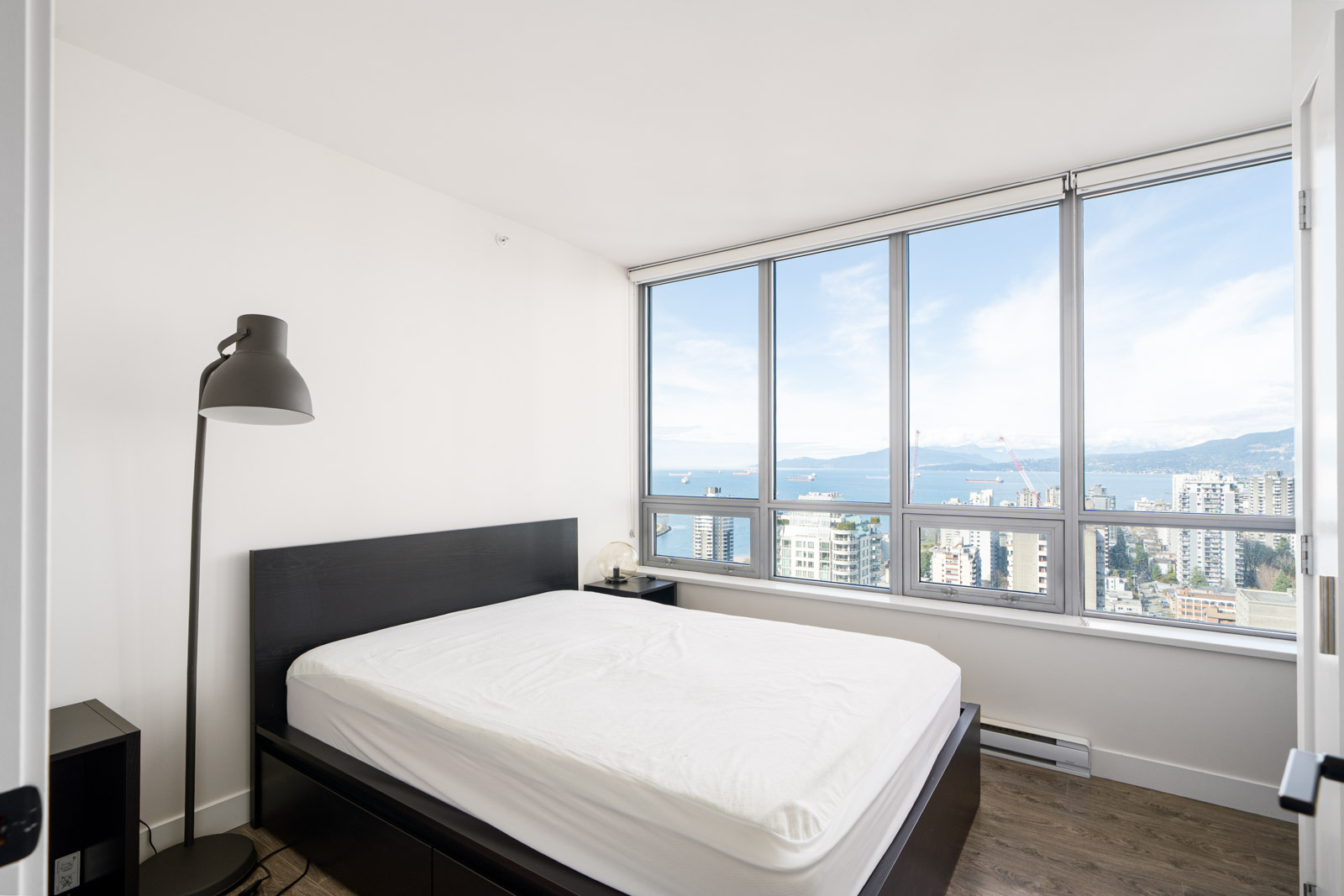 A minimal bedroom features a black bed frame, white mattress, and a tall floor lamp. Large windows reveal a cityscape and ocean view under a blue sky.