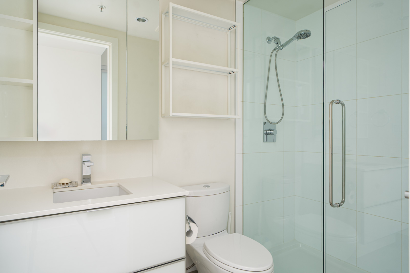 Modern bathroom with white fixtures, including a sink, toilet, and glass-enclosed shower. The room has a wall cabinet, mirrored cabinet, and a towel holder next to the toilet.