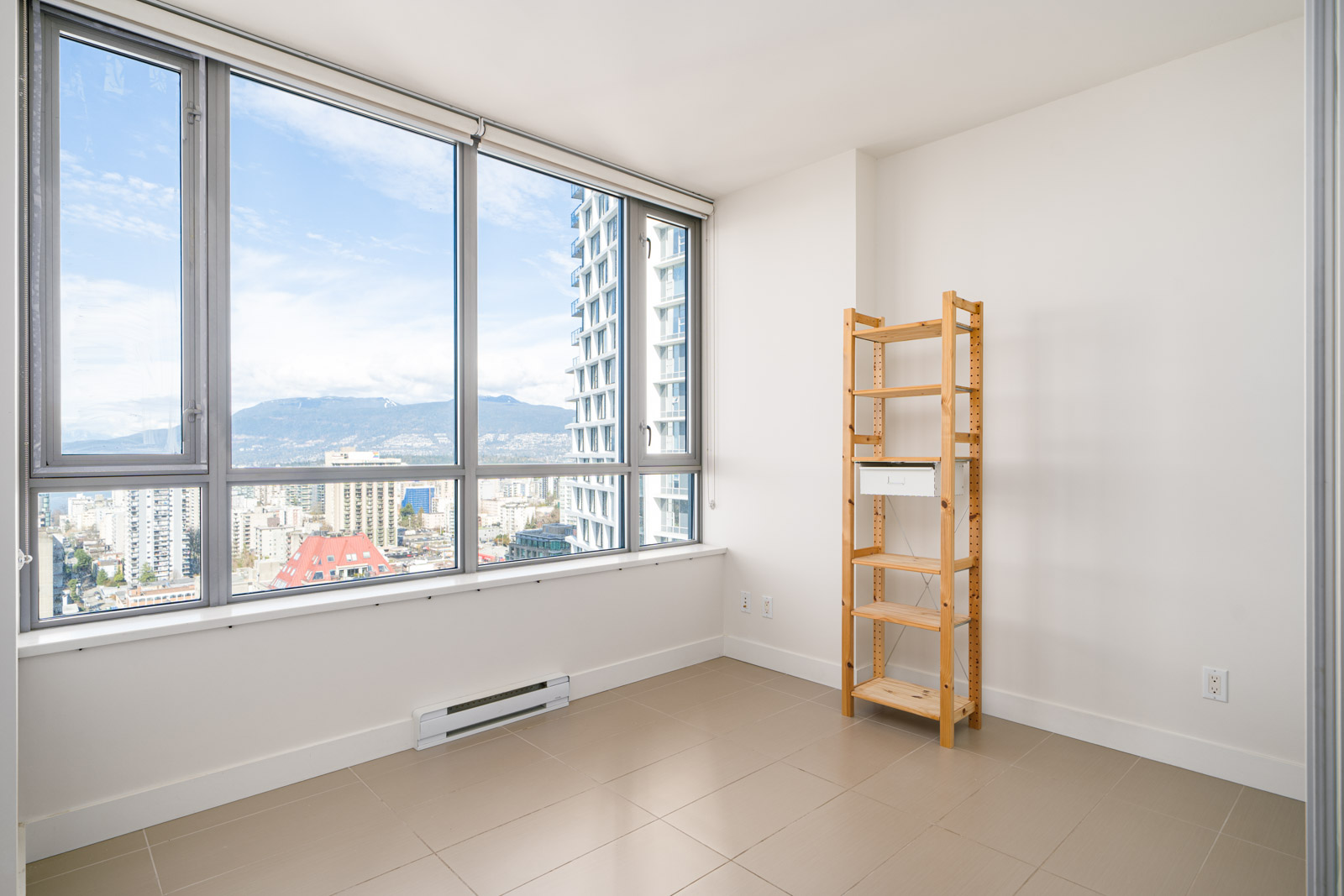Empty room with large windows showing a cityscape and mountains. A wooden shelving unit stands against a white wall. Tan tile flooring and a white baseboard heater are visible.