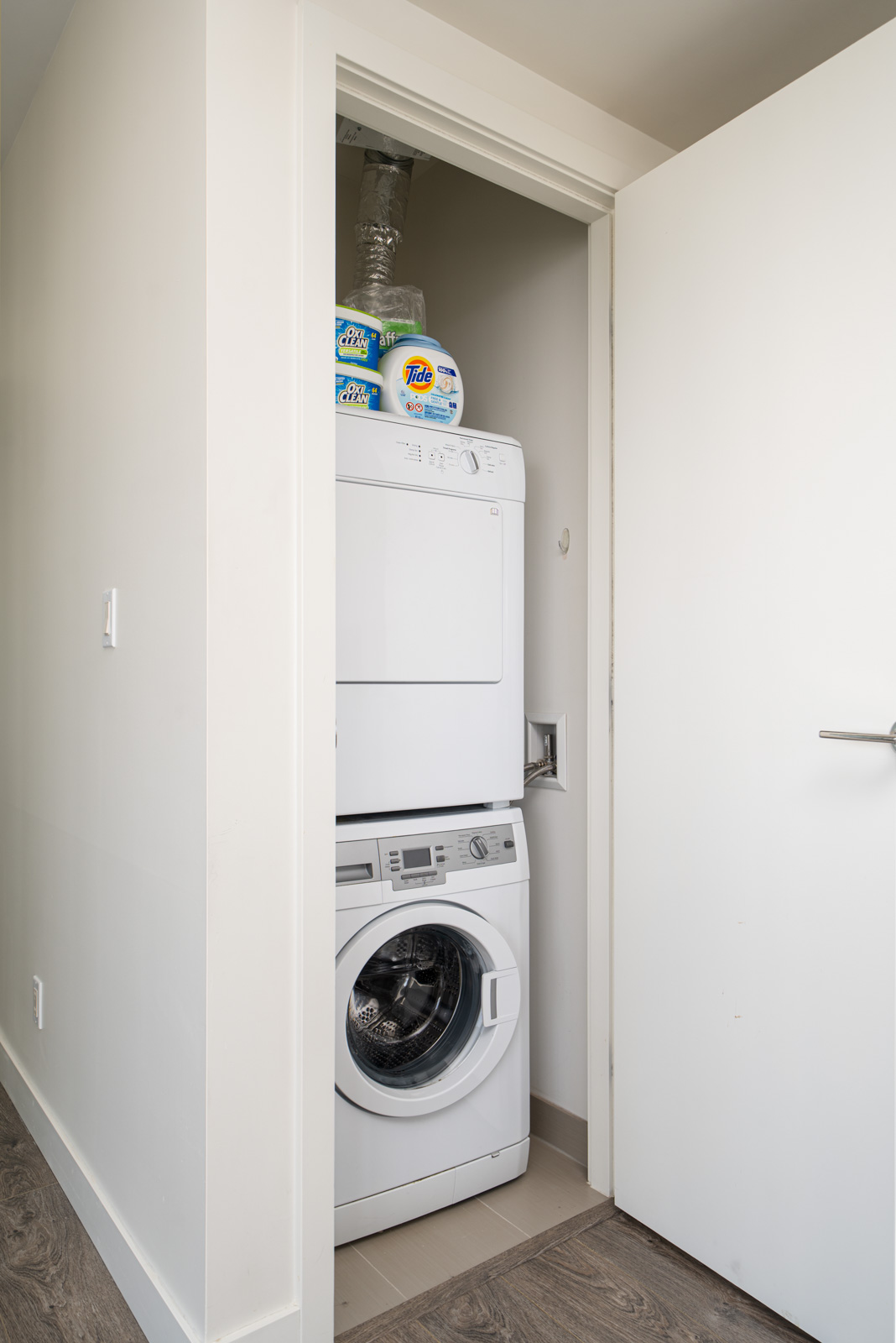 Stacked washer and dryer in a small laundry space with a partially open door, detergent bottles placed on top.
