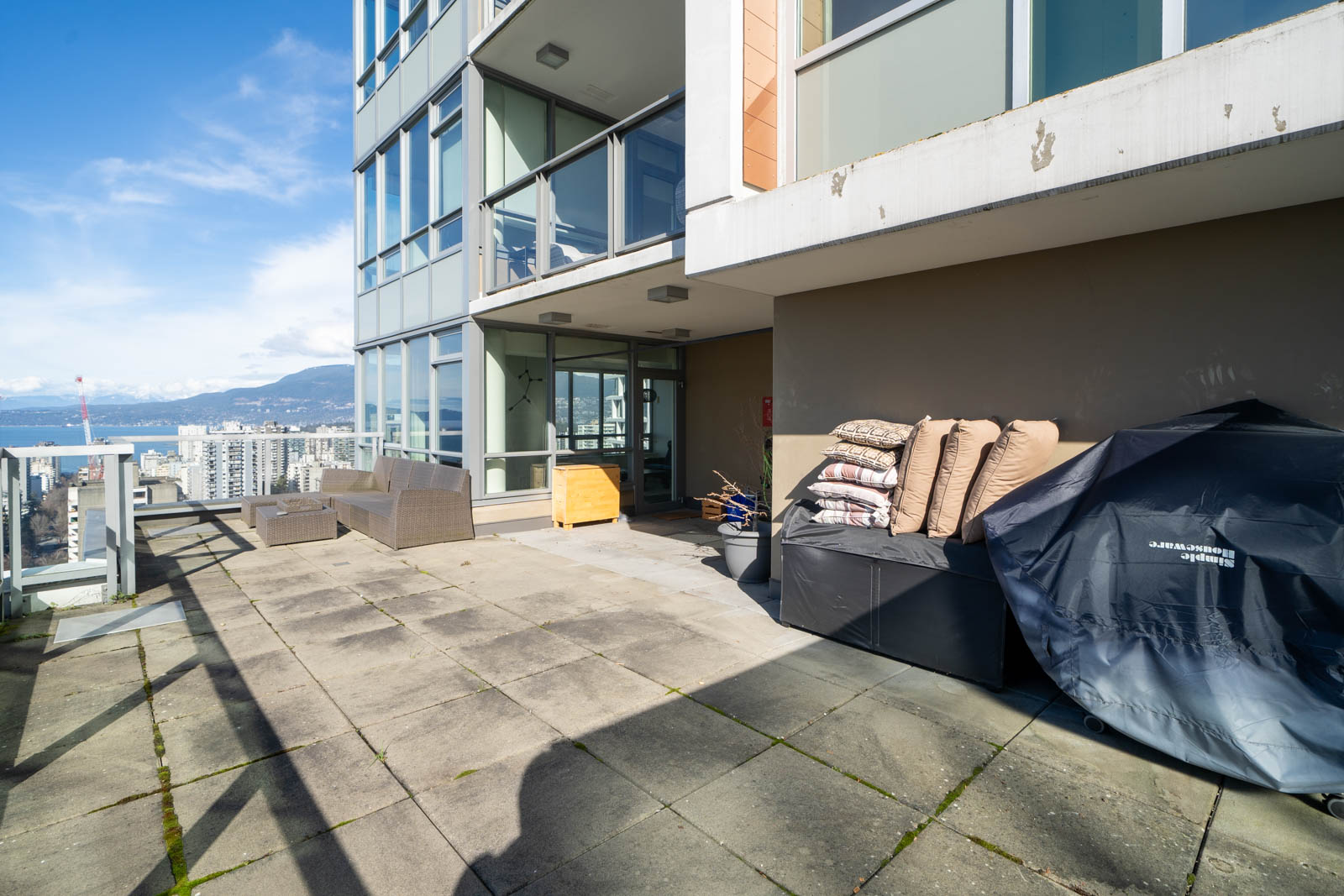 Rooftop patio with concrete tiles, outdoor furniture, covered grill, and stacked bags. Cityscape and mountain visible in the background under a clear sky.