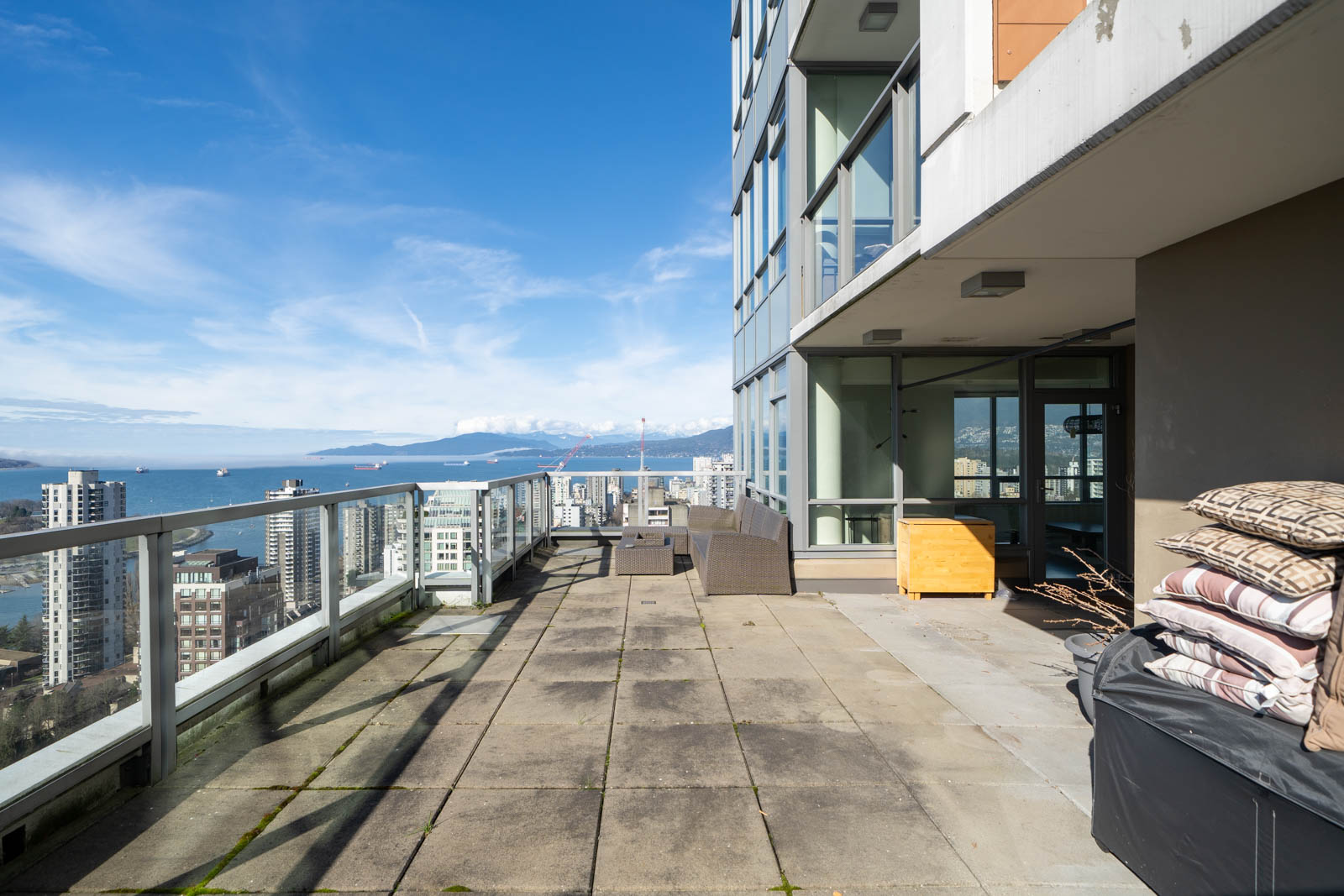 Rooftop terrace with glass railing, overlooking a cityscape and ocean. Outdoor furniture and stacked cushions are present.