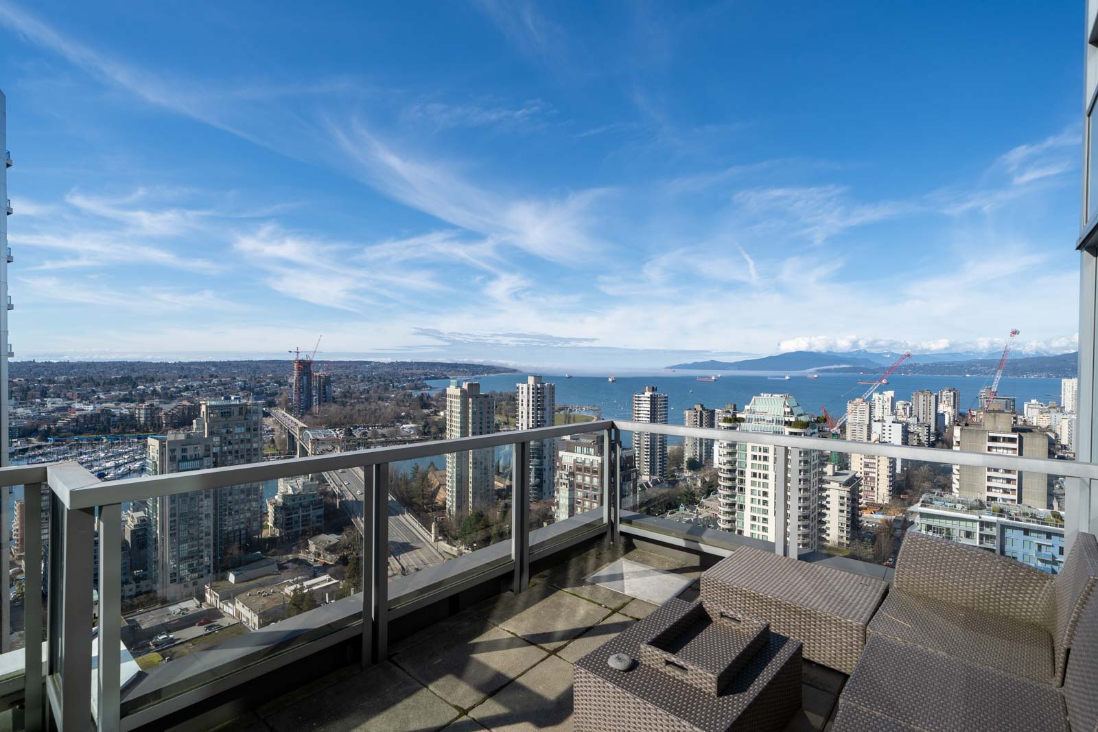 High-rise balcony view overlooking a cityscape with scattered buildings, a distant body of water, and mountains under a clear blue sky.