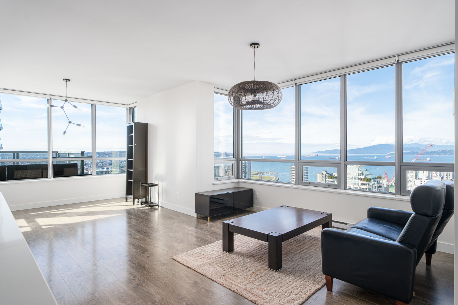 Modern living room with large windows offering a city and ocean view. Features a black armchair, coffee table, standing lamp, and decorative pendant light.