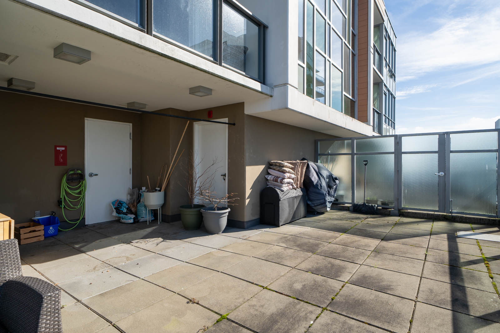 Outdoor patio with potted plants, a storage bin, hose, and broom. Glass partition on the right, and a modern building facade in the background.