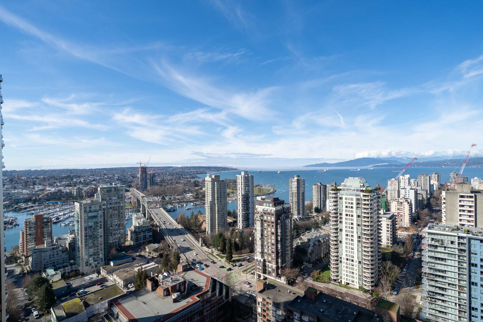 Aerial view of a coastal cityscape with high-rise buildings, a bridge, and a waterfront under a blue sky with scattered clouds.