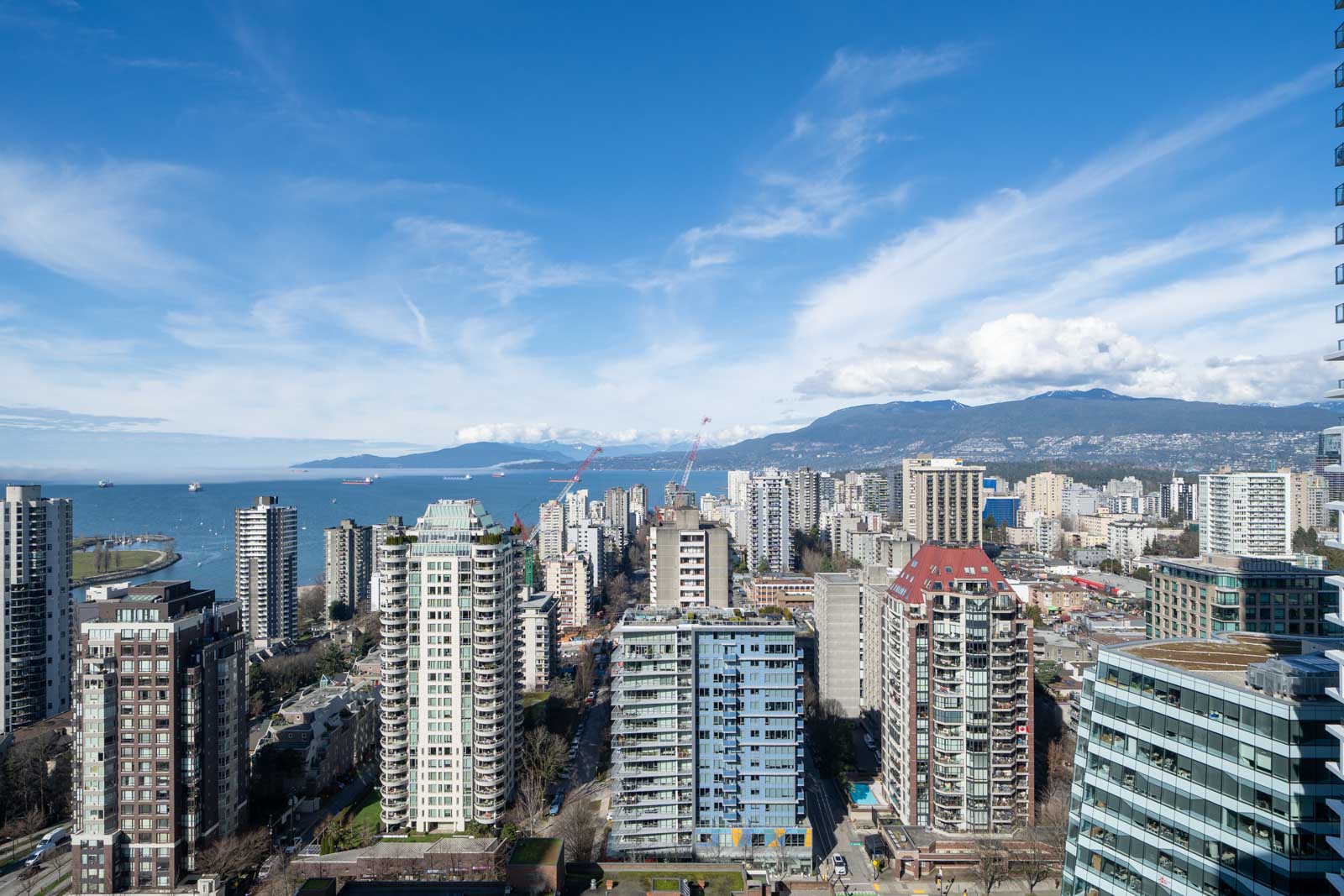 Aerial view of a city with numerous high-rise buildings, a body of water, and mountains in the background under a partly cloudy sky.