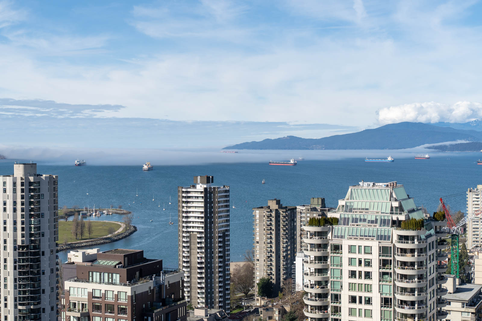 Aerial view of a cityscape with tall buildings along a waterfront. Several ships are in the bay, and mountains are visible in the distance under a partly cloudy sky.