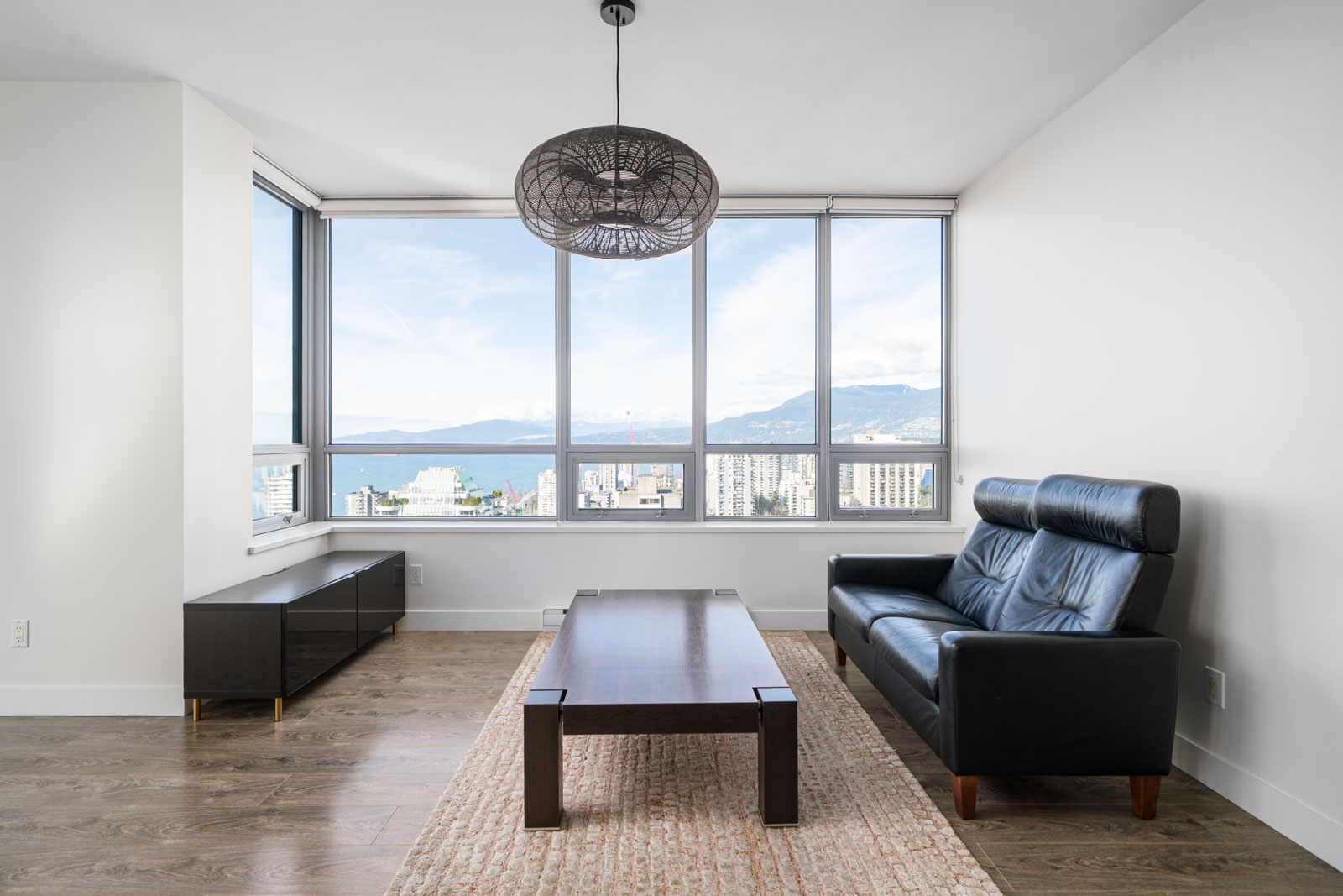 Modern living room with large windows, a black leather sofa, wooden coffee table, and sideboard. The windows offer a city skyline view with mountains in the background. Light fixture overhead.