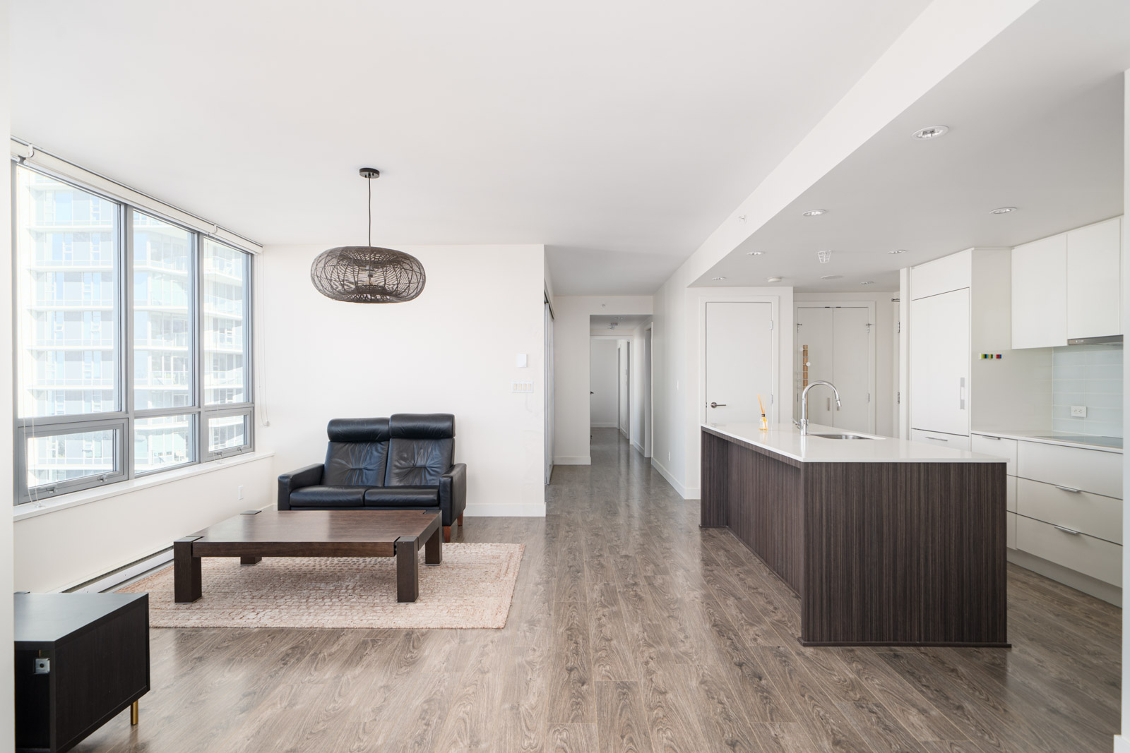 Modern open-plan apartment interior with a black sofa, wooden coffee table, kitchen island, and large windows. White walls and hardwood flooring create a bright, minimalist space.