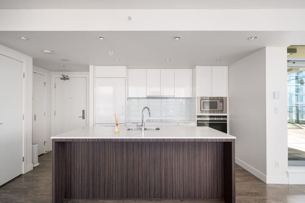 Modern kitchen with a large island, white cabinets, built-in oven, and a sleek faucet. Wooden floor and white walls enhance the minimalist design.