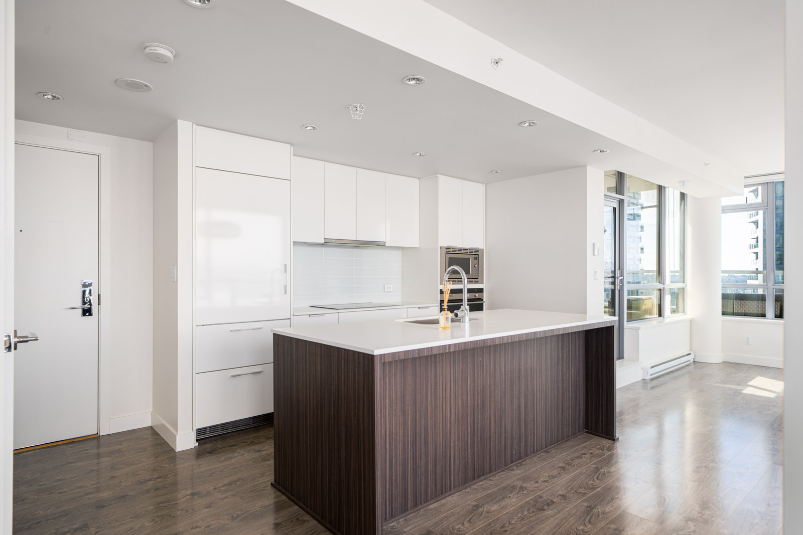 Modern kitchen with white cabinets, dark wood island, stainless steel appliances, and hardwood flooring. Large windows offer ample natural light. Minimalist design with open layout.