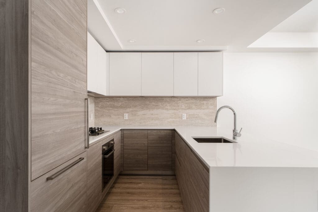 Modern kitchen with light wood cabinets, white countertops, and backsplash. Features built-in oven, gas stove, and sink under a bright, recessed ceiling.