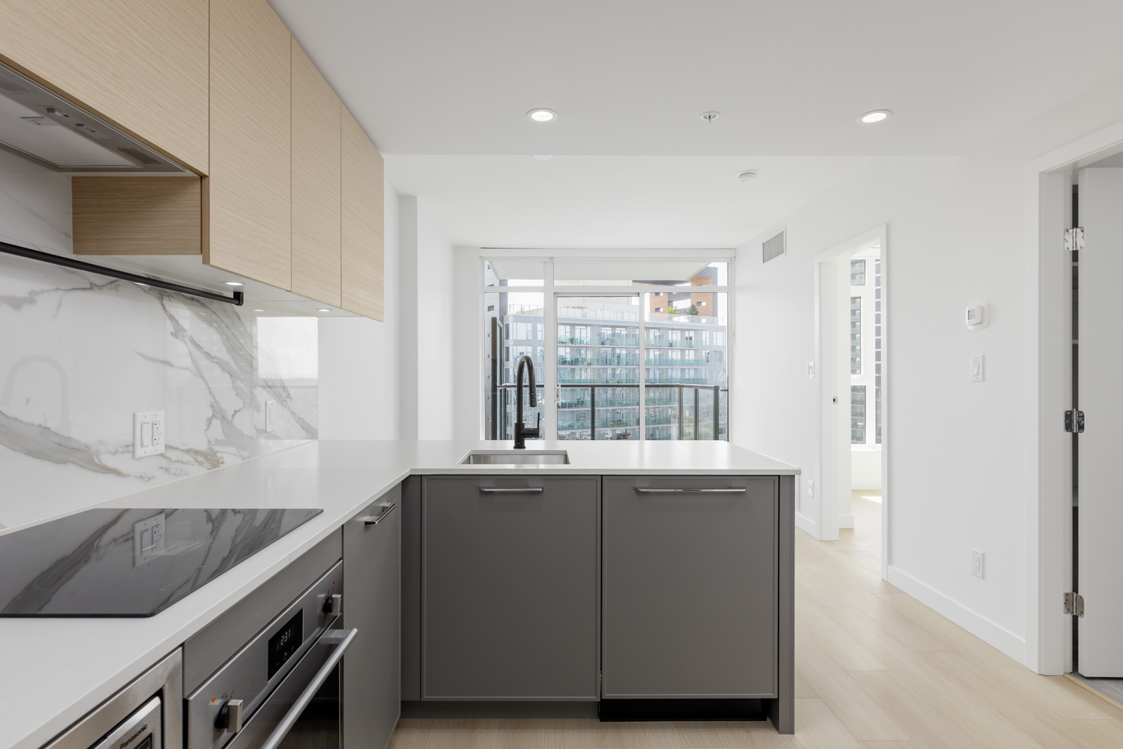 Modern kitchen with light wood cabinets, grey island with sink, marble backsplash, built-in appliances, and large windows overlooking a cityscape.