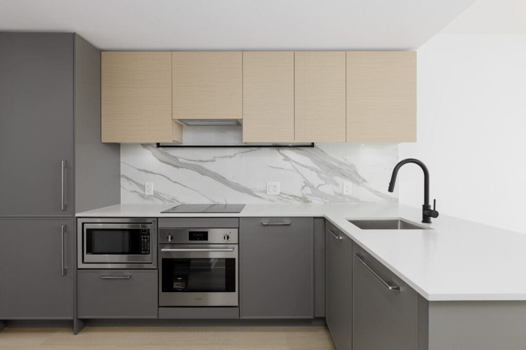 Modern kitchen with gray cabinets, built-in microwave and oven, electric cooktop, sink with black faucet, and white marble backsplash.