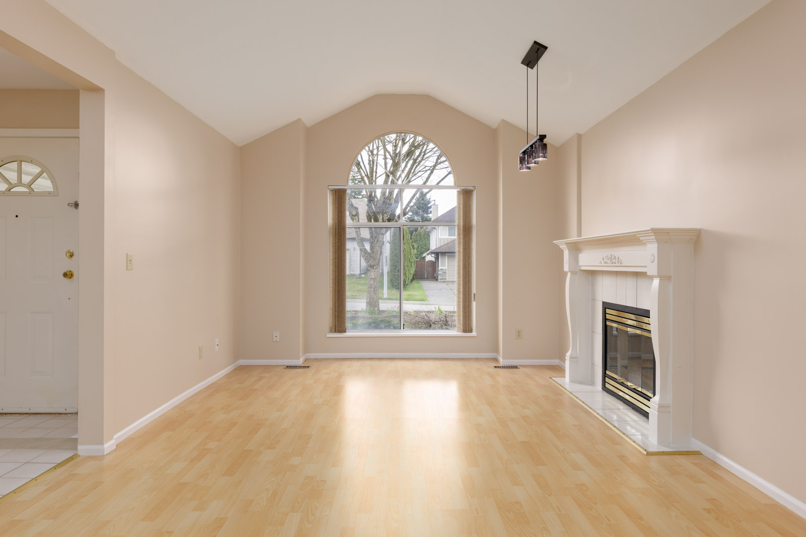 Empty living room with light wood flooring, beige walls, a large arched window, and a white fireplace on the right. A door is partially visible on the left.