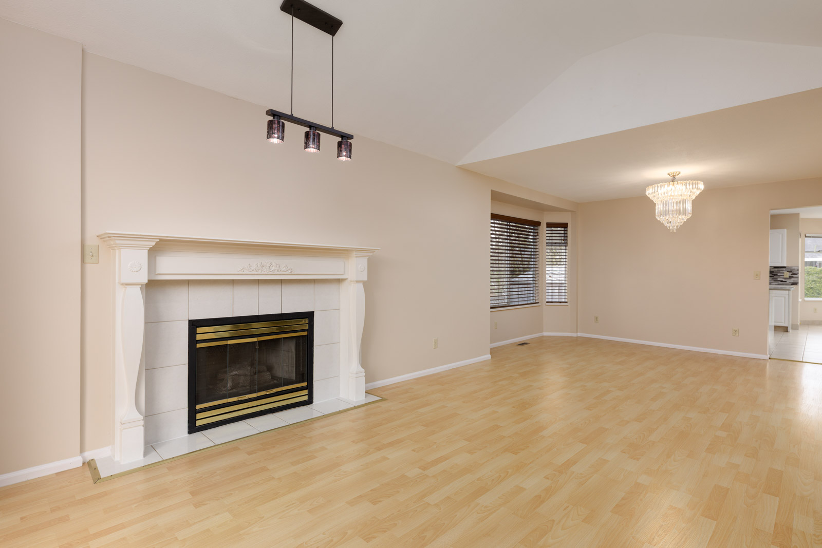 Empty living room with light wood flooring, a white fireplace on the left, a ceiling light fixture above, and a window with blinds on the right. An open doorway leads to another room.