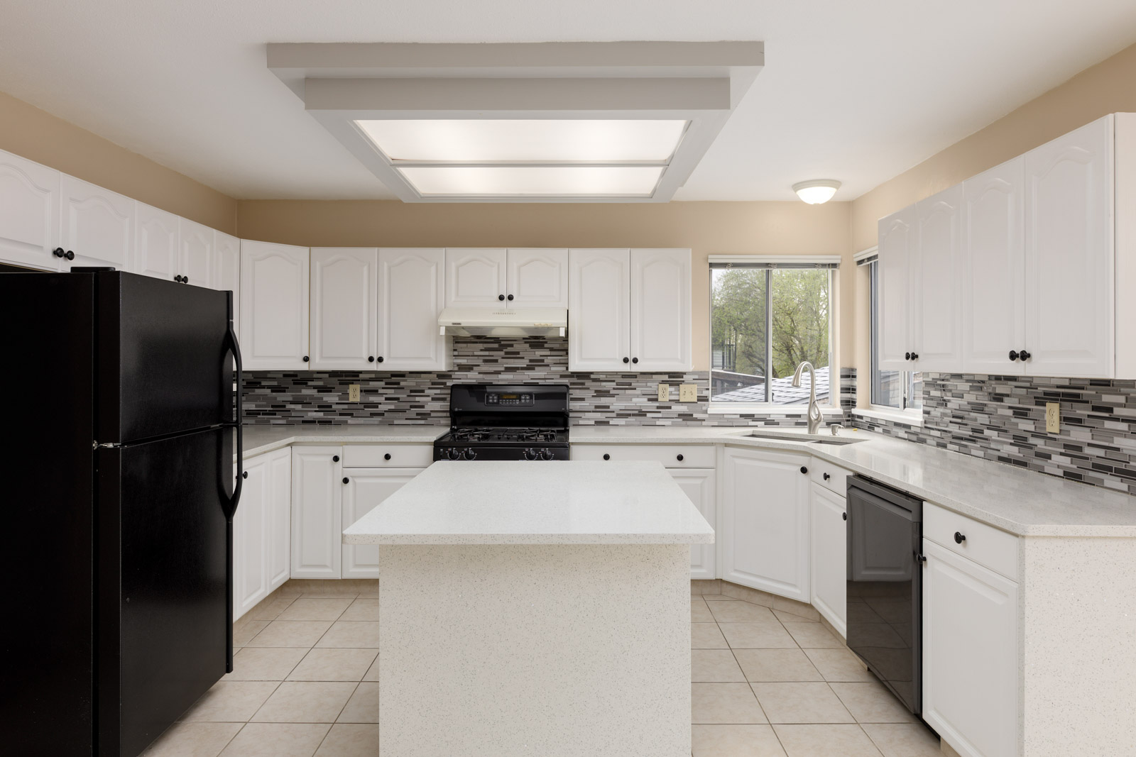 Modern kitchen with white cabinets, black appliances, and a central island. Features tiled backsplash, large overhead light, and tiled floor. Window with a view of greenery outside.