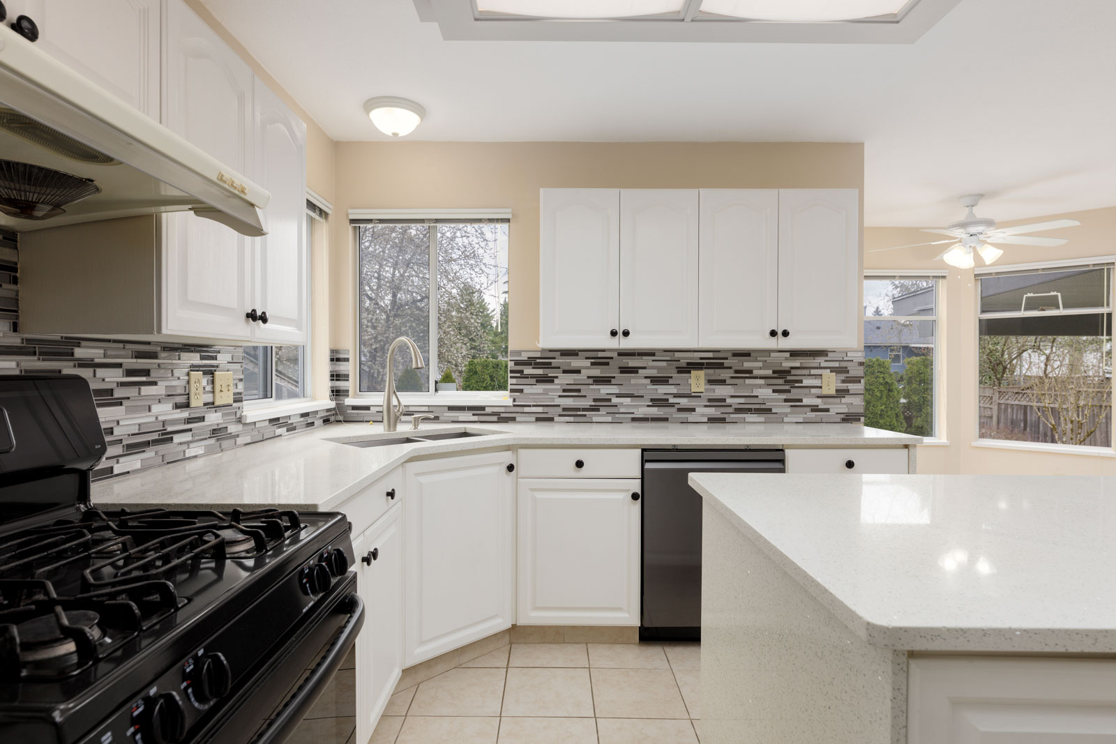 Modern kitchen with white cabinets, black appliances, tiled backsplash, and large windows. Features a gas stove and central island with light-colored countertops.