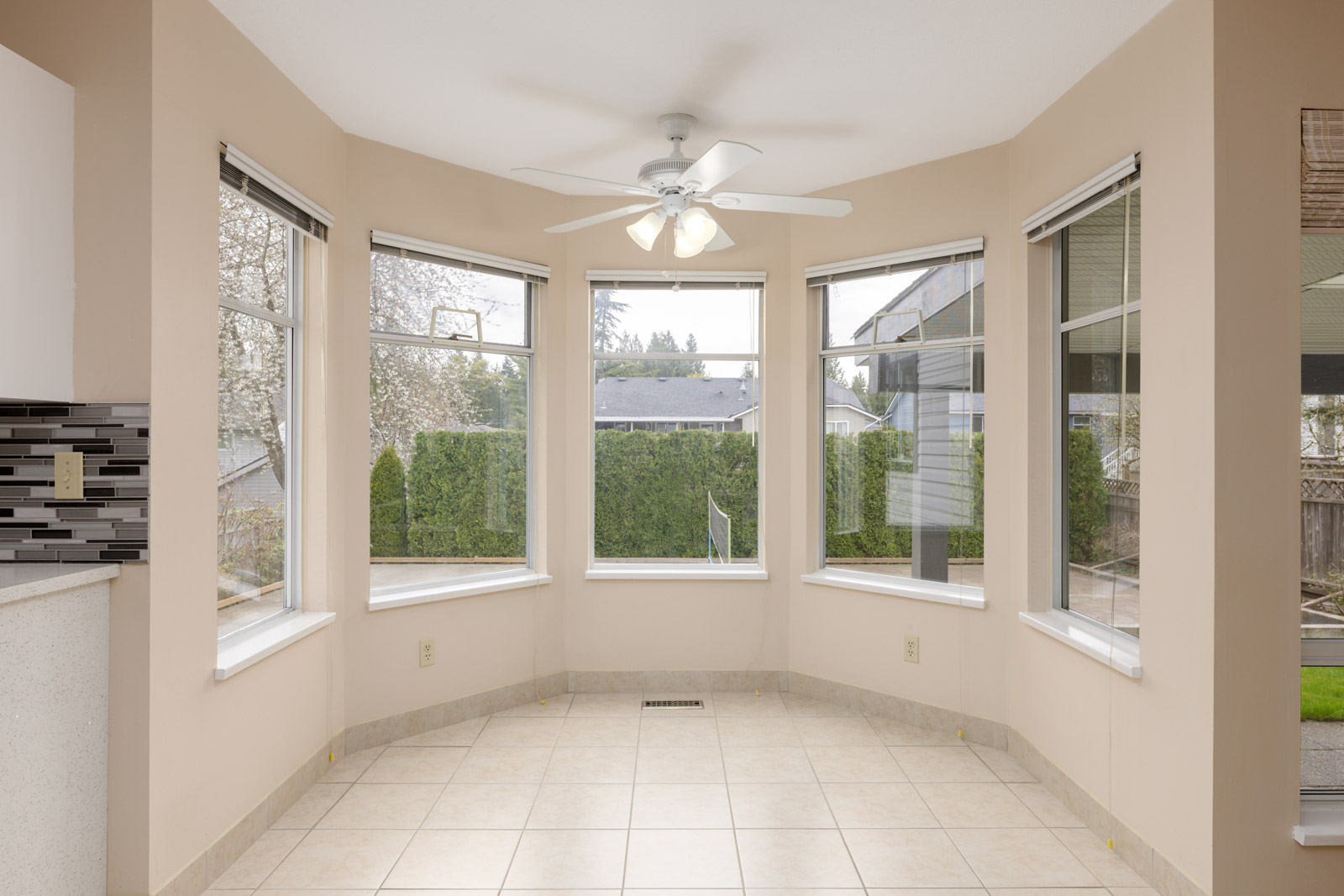 A sunlit room with five large bay windows, tiled floor, beige walls, and a ceiling fan, overlooking a fenced backyard with tall hedges.