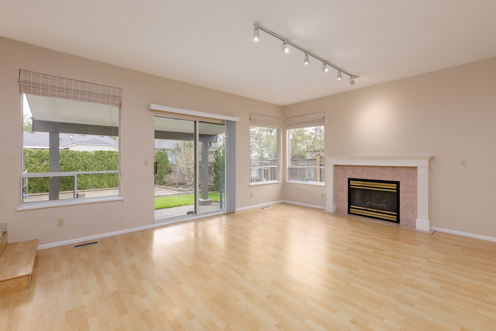 Spacious, empty living room with light wooden flooring, a glass sliding door to a patio, windows, track lighting, and a fireplace with a mantel.