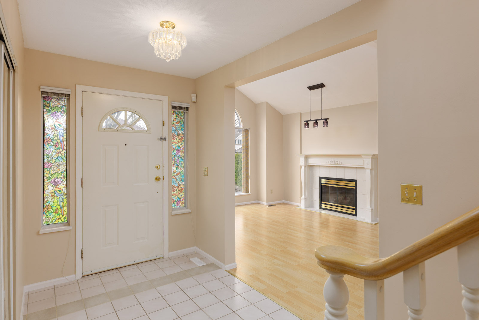 Home entryway with a white door framed by stained glass panels. Adjacent room features a fireplace, light-colored hardwood flooring, and a modern light fixture above.