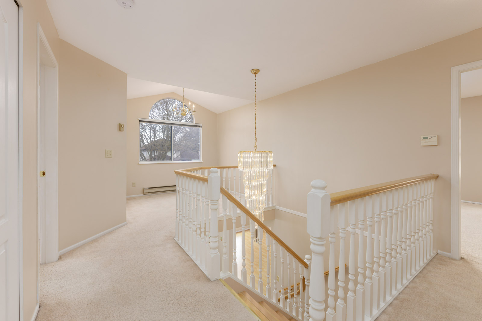 Cream-colored carpeted hallway with a central staircase featuring a chandelier. There are two doorways and a window with a view of trees across the room.
