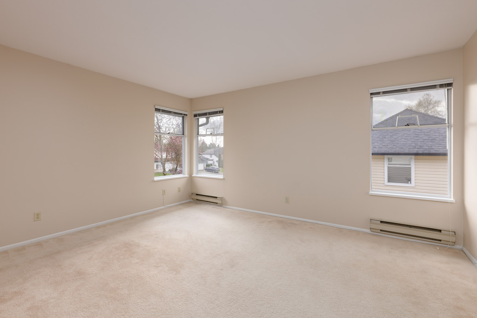 An empty beige-walled room with two windows, beige carpet flooring, and baseboard heaters beneath the windows.