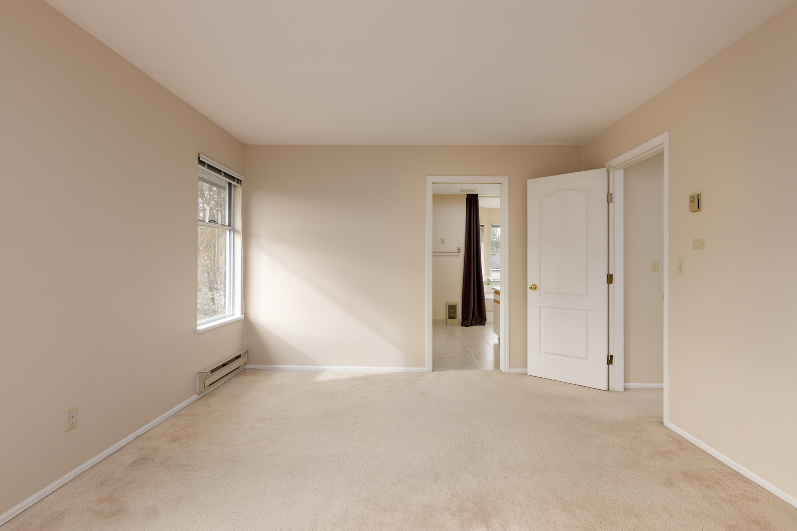 Empty beige room with carpet, a window on the left, open door leading to another room, wall heater, and light switch.