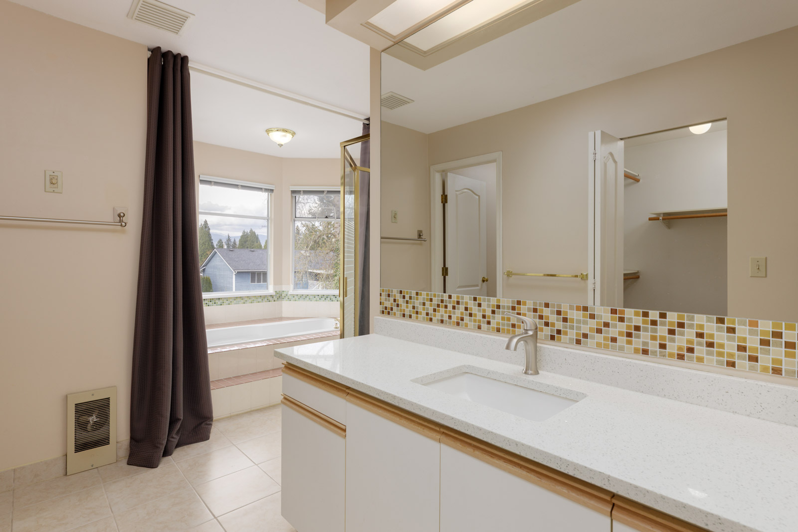 Bathroom with a large mirror, white countertop, mosaic tile backsplash, and white sink. Bathtub near a window with dark curtains. Shelves and a door in the background.
