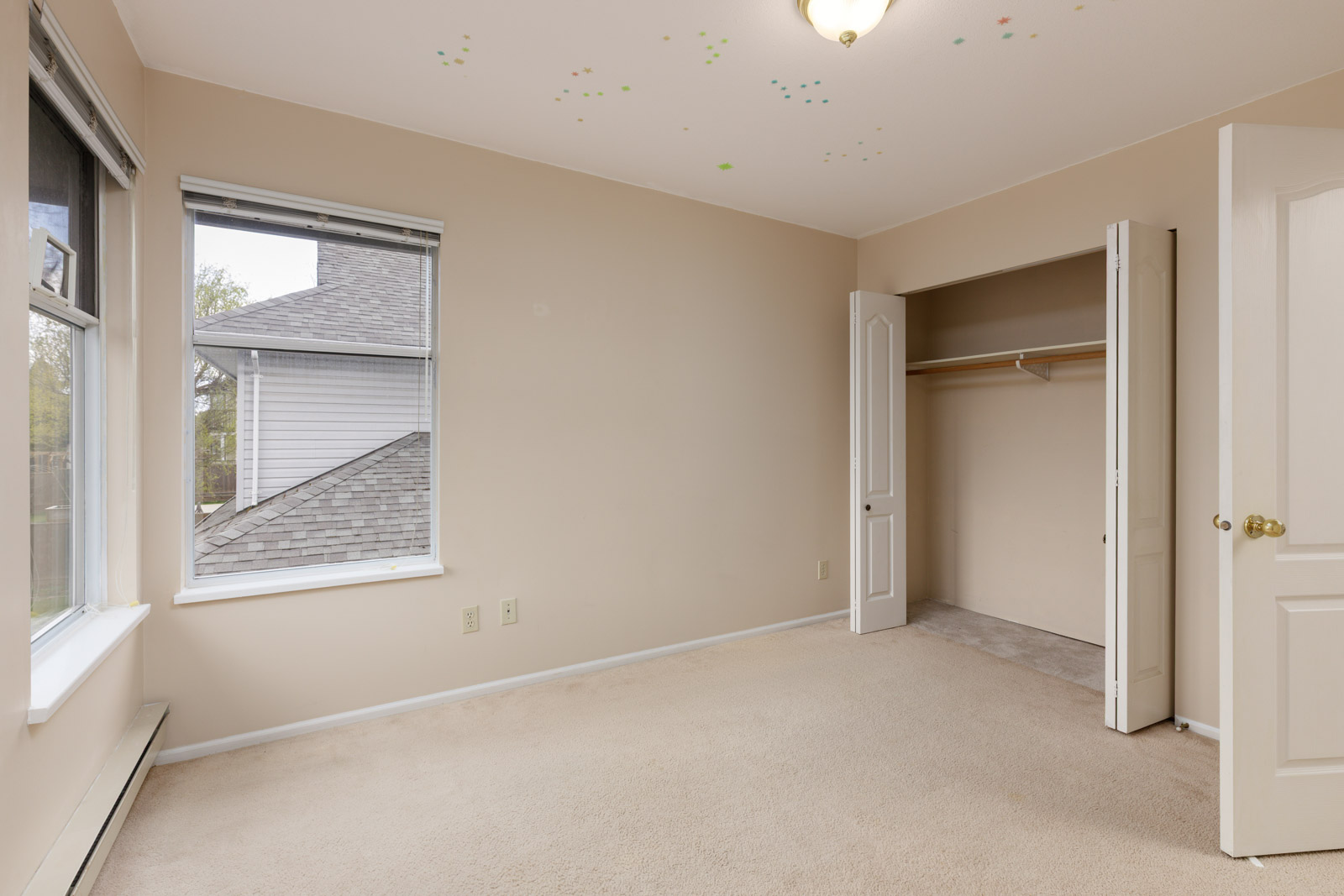 Empty beige room with a closet door open, two windows showing a roof view, and ceiling light fixture. Colorful stickers are on the ceiling. Carpeted floor.